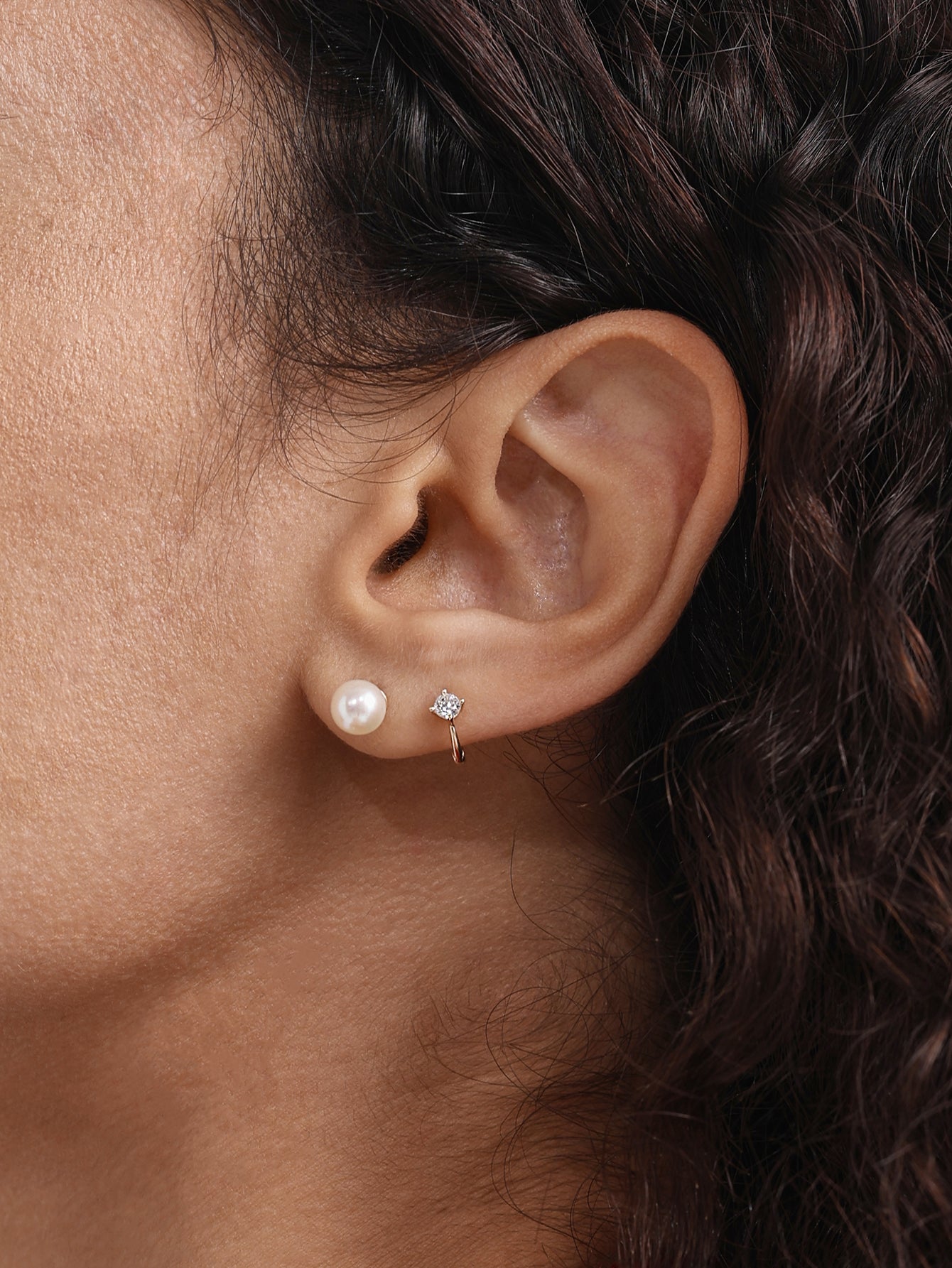 14K solid gold mini huggie hoop earring with a sparkling cubic zirconia, shown on earlobe next to a pearl stud.