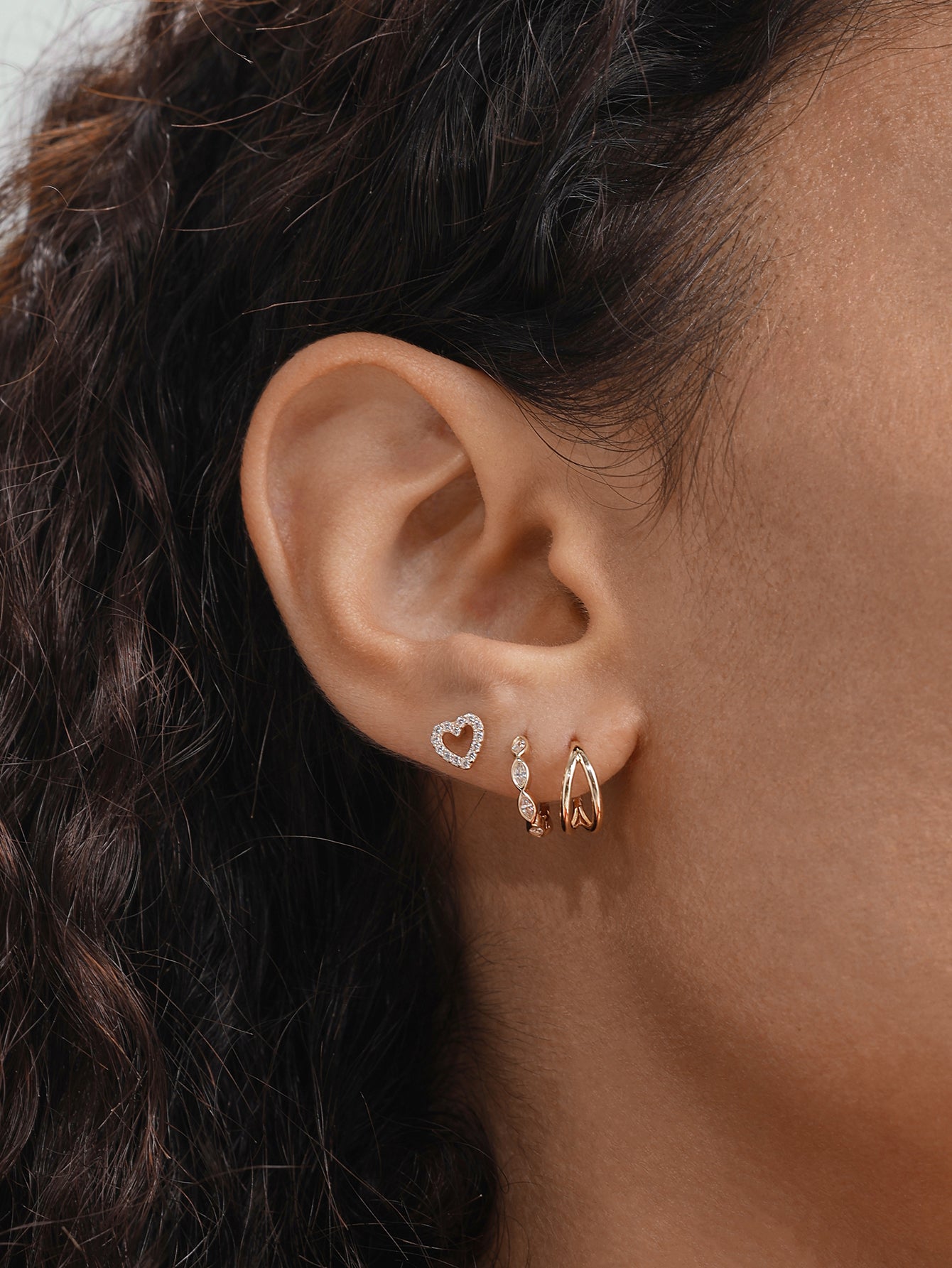 Woman's ear featuring a 14K solid gold crystal hinged hoop clicker, alongside a heart stud and linear drop earring.