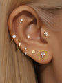 14K solid gold lucky cross ear piercing stud with cubic zirconia on human ear, shown with other gold earrings.