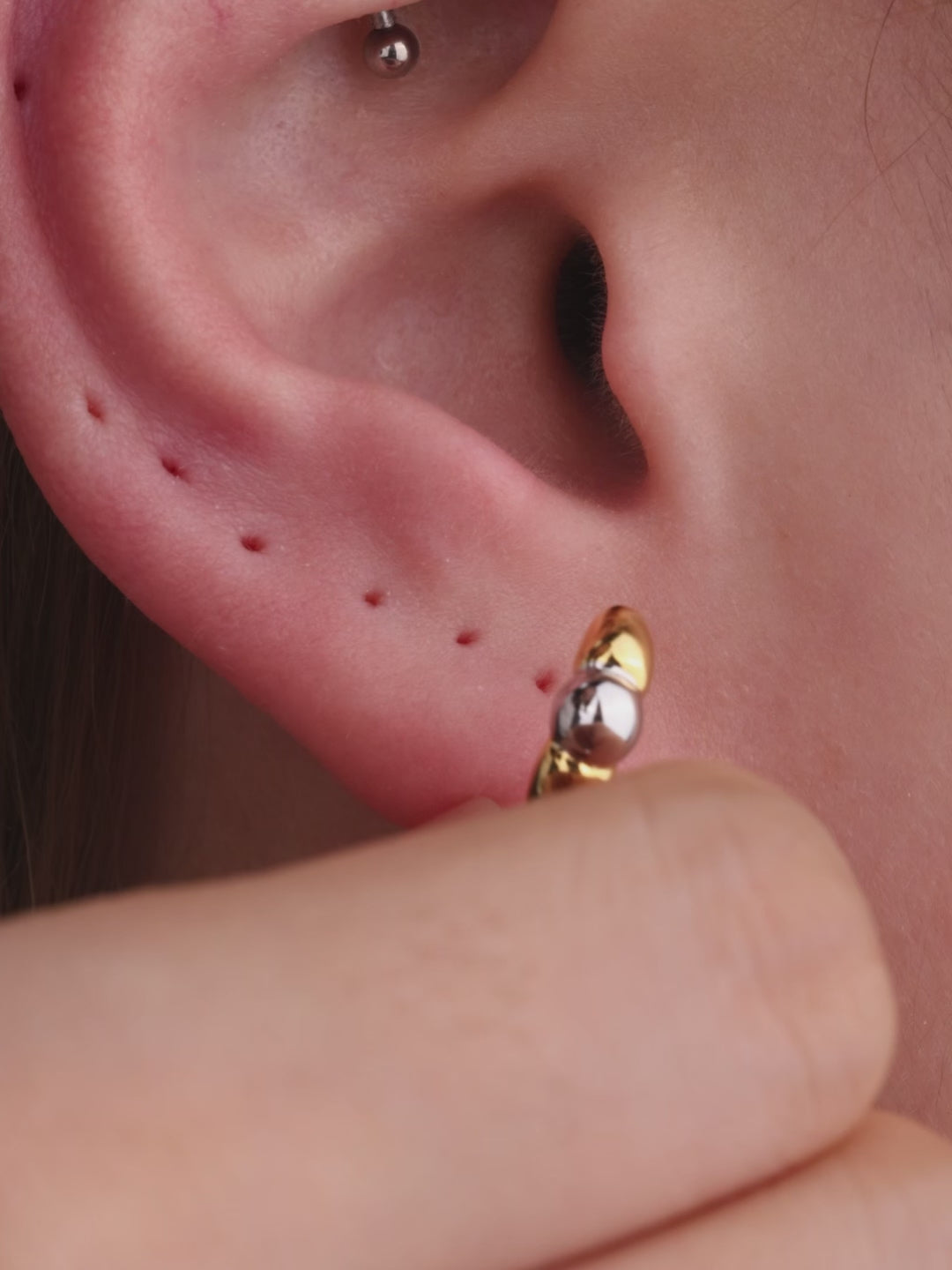 Sophisticated layered ear styling showcasing KOSINER Perléa beaded cross stud connected with delicate chain to teardrop outline piercing, paired with north star helix studs, marquise detail daith clicker, blue marquise eye stud and two-tone huggie hoop, trendy versatile curated ear layout
